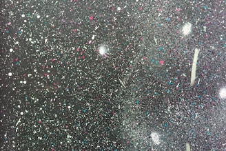 A textured surface covered in colorful splatters of paint, featuring a mix of white, blue, purple, and pink against a dark background. The paint droplets vary in size and form abstract patterns, giving the appearance of a starry night sky or a cosmic scene.