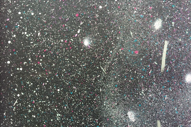 A textured surface covered in colorful splatters of paint, featuring a mix of white, blue, purple, and pink against a dark background. The paint droplets vary in size and form abstract patterns, giving the appearance of a starry night sky or a cosmic scene.