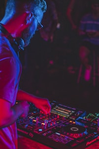 A person is focused on operating DJ equipment, surrounded by an array of controls, buttons, and lights. The atmosphere is dimly lit with vibrant, colorful lighting accenting the DJ setup, creating a lively and immersive ambiance.
