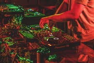 A person is mixing music on professional DJ equipment, surrounded by vibrant red and green lighting. The setup is on a wooden table adorned with some leaves, creating a lively atmosphere. The DJ's hands are actively manipulating the controls.