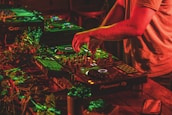 A person is mixing music on professional DJ equipment, surrounded by vibrant red and green lighting. The setup is on a wooden table adorned with some leaves, creating a lively atmosphere. The DJ's hands are actively manipulating the controls.