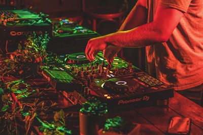 A person is mixing music on professional DJ equipment, surrounded by vibrant red and green lighting. The setup is on a wooden table adorned with some leaves, creating a lively atmosphere. The DJ's hands are actively manipulating the controls.