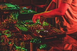A person is mixing music on professional DJ equipment, surrounded by vibrant red and green lighting. The setup is on a wooden table adorned with some leaves, creating a lively atmosphere. The DJ's hands are actively manipulating the controls.