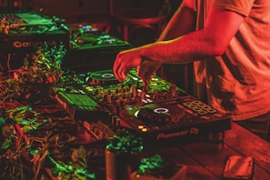 A person is mixing music on professional DJ equipment, surrounded by vibrant red and green lighting. The setup is on a wooden table adorned with some leaves, creating a lively atmosphere. The DJ's hands are actively manipulating the controls.
