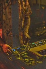 A person is wearing a colorful floral shirt and is using a DJ controller. The dim lighting creates a warm, intimate atmosphere. Vines or plant-like decorations are placed on the equipment, adding an organic element to the scene.