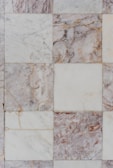 Square tiles made of marble with varying shades of white, gray, and brown. Some squares display intricate veins of darker and lighter colors, giving a textured and natural appearance. The tiles are arranged in a grid pattern, with visible seams and occasional signs of wear or imperfections.