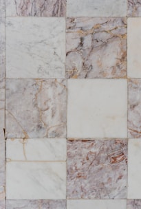 Square tiles made of marble with varying shades of white, gray, and brown. Some squares display intricate veins of darker and lighter colors, giving a textured and natural appearance. The tiles are arranged in a grid pattern, with visible seams and occasional signs of wear or imperfections.
