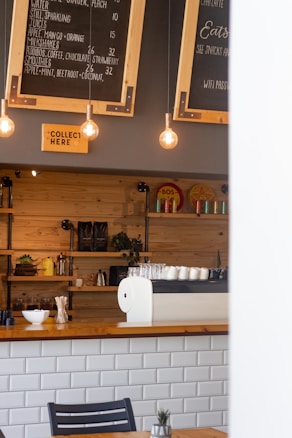 A cozy cafe interior with a wooden counter, a white tiled front, and a black chair partially visible in the foreground. Above the counter, there are hanging light bulbs and blackboards displaying the menu, including juices and smoothies. Shelves behind the counter hold various items such as cups, decorative objects, and coffee supplies. A sign that reads 'Collect Here' is also visible.