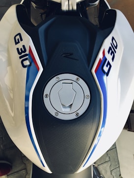 A motorcycle fuel tank with a silver fuel cap and branding that reads 'G 310' in blue with a red and blue stripe. The tank is predominantly black with white and blue accents.