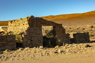 An ancient stone structure partially intact sits in a barren desert landscape, with stacks of weathered stones forming its walls. Sparse vegetation grows around the ruins, and the ground is littered with rocks. The backdrop features a barren hillside under clear blue skies.
