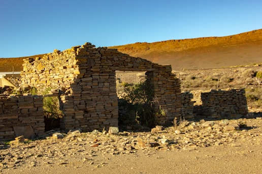An ancient stone structure partially intact sits in a barren desert landscape, with stacks of weathered stones forming its walls. Sparse vegetation grows around the ruins, and the ground is littered with rocks. The backdrop features a barren hillside under clear blue skies.