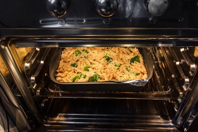 An oven with the door open reveals a rectangular baking dish inside. The dish contains a pasta bake with penne, broccoli, and a covering of melted cheese. The oven's interior is illuminated and clean, with a shiny metallic surface.