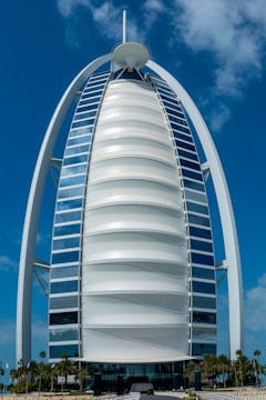 A sleek, modern sail-shaped skyscraper with reflective blue and white panels, set against a bright blue sky with scattered clouds. Palm trees line the foreground, adding a touch of greenery.