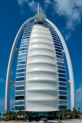 A sleek, modern sail-shaped skyscraper with reflective blue and white panels, set against a bright blue sky with scattered clouds. Palm trees line the foreground, adding a touch of greenery.