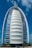 A sleek, modern sail-shaped skyscraper with reflective blue and white panels, set against a bright blue sky with scattered clouds. Palm trees line the foreground, adding a touch of greenery.