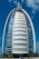 A sleek, modern sail-shaped skyscraper with reflective blue and white panels, set against a bright blue sky with scattered clouds. Palm trees line the foreground, adding a touch of greenery.