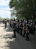A group of uniformed band members standing in formation on a shaded street. They are wearing dark, formal military-style uniforms with shiny helmets. Some hold musical instruments, such as saxophones and trumpets. In the background, a few onlookers and trees are visible, adding depth to the scene.