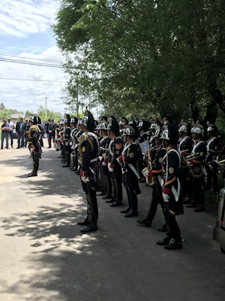 A group of uniformed band members standing in formation on a shaded street. They are wearing dark, formal military-style uniforms with shiny helmets. Some hold musical instruments, such as saxophones and trumpets. In the background, a few onlookers and trees are visible, adding depth to the scene.