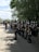 A group of uniformed band members standing in formation on a shaded street. They are wearing dark, formal military-style uniforms with shiny helmets. Some hold musical instruments, such as saxophones and trumpets. In the background, a few onlookers and trees are visible, adding depth to the scene.