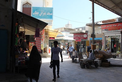 A busy marketplace scene with several people walking and sitting. Stalls are filled with goods, and various signs in a foreign script are visible. Buildings have a mix of traditional and modern architecture. The lighting suggests it is daytime.