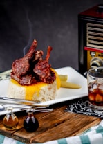 A beautifully plated dish features glossy, succulent lamb chops atop a mound of fluffy white rice with a golden sauce drizzle. The wooden table setting includes a glass of dark soda with ice, small bottles of condiments, and cutlery. In the background, there is a vintage-style radio adding a retro touch.