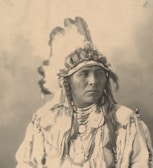 A person wearing a traditional Native American headdress adorned with feathers and beads. The attire includes necklaces and clothing with intricate patterns. The image is in sepia tones, providing a historical or vintage feel.