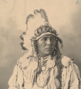 A person wearing a traditional Native American headdress adorned with feathers and beads. The attire includes necklaces and clothing with intricate patterns. The image is in sepia tones, providing a historical or vintage feel.