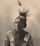 A sepia-toned portrait of an elderly person wearing traditional attire with intricate patterns. The hair is styled with a feather, and the expression is thoughtful and serene.