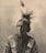 A sepia-toned portrait of an elderly person wearing traditional attire with intricate patterns. The hair is styled with a feather, and the expression is thoughtful and serene.