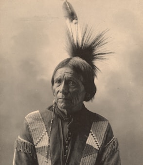 A sepia-toned portrait of an elderly person wearing traditional attire with intricate patterns. The hair is styled with a feather, and the expression is thoughtful and serene.