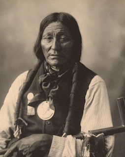 An older person with long hair seated in a studio setting. They wear a dark vest over a light-colored shirt, with a large medallion hanging around their neck. Their expression is calm and introspective, and they hold a long pipe with decorative elements.
