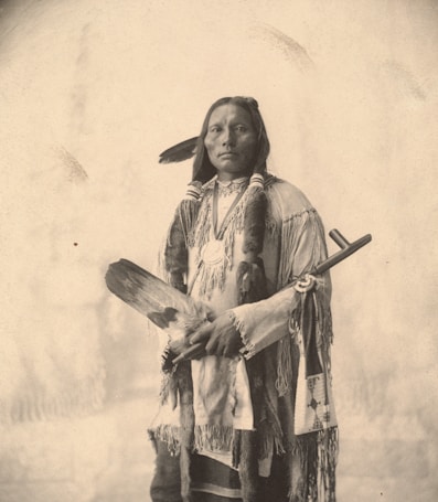 A person dressed in traditional indigenous attire with long braids and feathers stands against a plain background. They hold a feathered object in their hands and wear intricate jewelry and garments adorned with patterns.