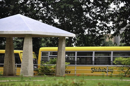 A yellow school bus is partially visible with its distinctive coloring parked in a lush, green environment. A concrete shelter with a triangular roof is in the foreground, surrounded by greenery and trees, offering a contrast between man-made and natural elements.