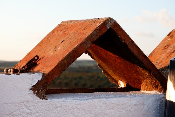 A close-up of a terracotta roof tile structure in a triangular shape, with some metal components attached. The background shows a blurred view of a natural landscape under a clear sky.