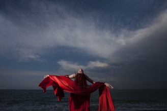 woman in red dress standing on sea shore under gray cloudy sky during daytime