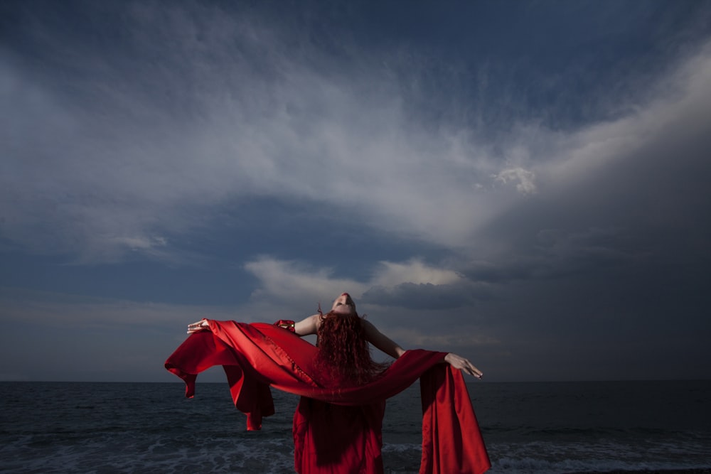 Frau in rotem Kleid, die tagsüber am Meeresufer unter grauem, bewölktem Himmel steht