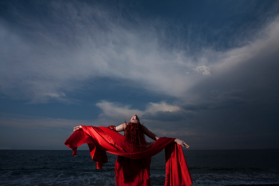 woman in red dress standing on sea shore under gray cloudy sky during daytime