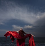 woman in red dress standing on sea shore under gray cloudy sky during daytime