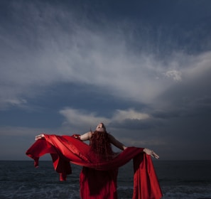 woman in red dress standing on sea shore under gray cloudy sky during daytime