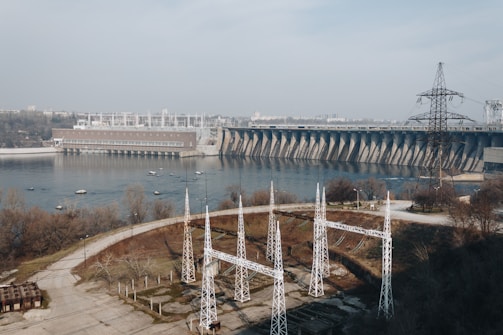 A large hydroelectric dam spans across a wide river, with utility poles and electrical infrastructure in the foreground. The surrounding area features sparse vegetation and paved roads. In the distance, there are buildings indicating an urban setting.