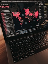 A laptop displays a global map visualization of COVID-19 cases, showing data from various countries. The left panel lists confirmed cases by country, with China having the highest count. The right panel displays the total number of deaths and recovered cases. Circles of varying sizes on the map indicate the concentration of cases in different regions.