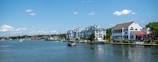 A waterfront scene featuring several modern residential buildings along the edge of a calm body of water. Several boats are moored near the shore, with one in the foreground and others in the distance. The sky is clear and blue with a few scattered clouds.
