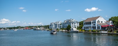 A waterfront scene featuring several modern residential buildings along the edge of a calm body of water. Several boats are moored near the shore, with one in the foreground and others in the distance. The sky is clear and blue with a few scattered clouds.