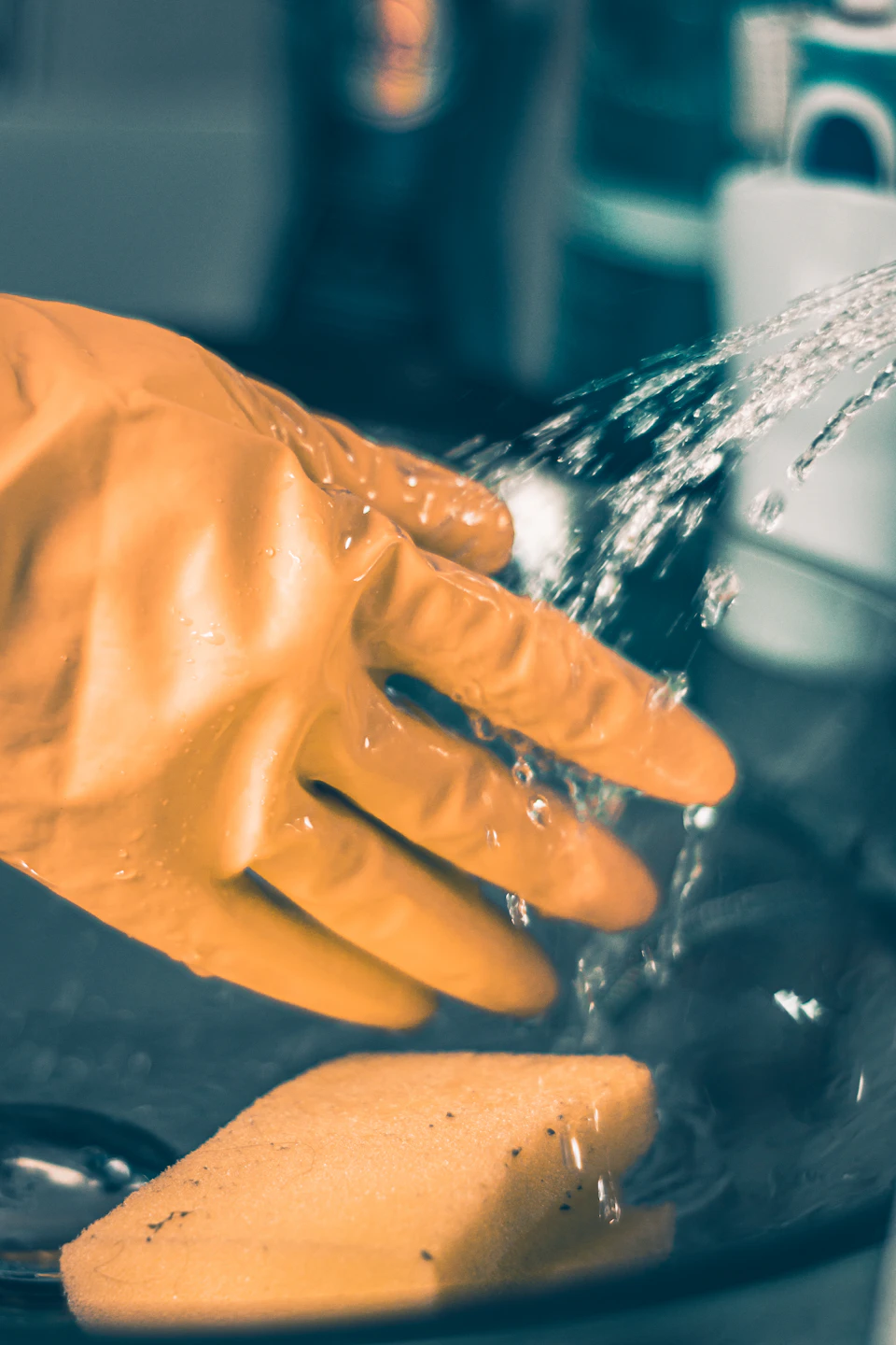 Person's hand wears orange rubber glove while washing dishes with sponge under running water