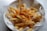 A plate of crispy golden french fries topped with melted cheese and garnished with herbs. The fries are served in a white bowl lined with branded paper.