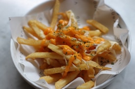 A plate of crispy golden french fries topped with melted cheese and garnished with herbs. The fries are served in a white bowl lined with branded paper.