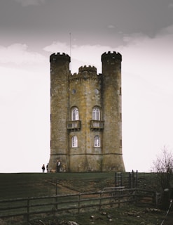 A stone tower with two round turrets stands prominently against a cloudy sky. The structure features arched windows and a central area connecting the turrets. In the foreground, a few people and a dog are visible near the base of the tower, with a wooden fence and some sparse vegetation on grassy terrain.