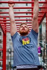 A man is performing an exercise on a set of red monkey bars outdoors. He is reaching up to hold onto one of the bars, wearing a gray t-shirt with a logo and text on it. The background features a blurred image of urban architecture.