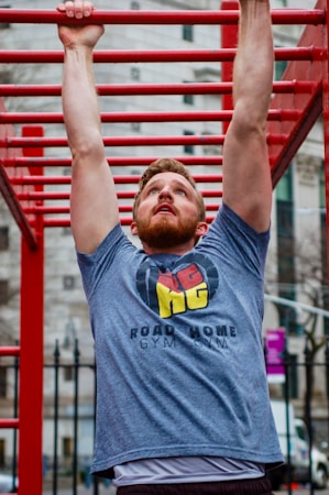 A man is performing an exercise on a set of red monkey bars outdoors. He is reaching up to hold onto one of the bars, wearing a gray t-shirt with a logo and text on it. The background features a blurred image of urban architecture.