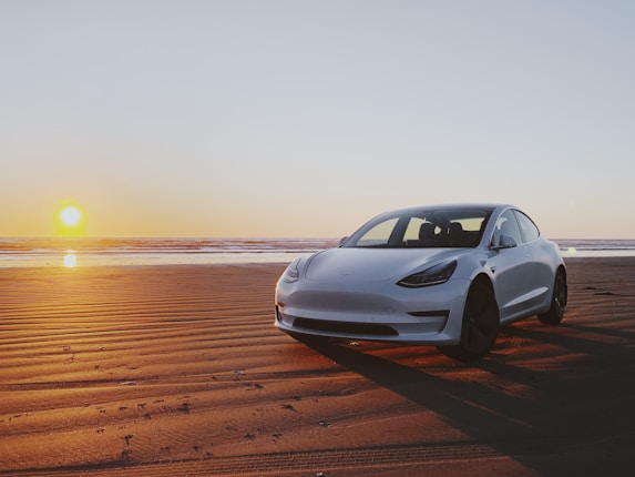 A sleek white car parked on a sandy beach during sunset, with the sun low on the horizon casting a warm glow over the scenery. The sky has a smooth gradient from blue to orange, and gentle waves are visible in the background.